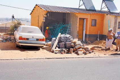 Gogo Nomgqibelo Mkhwanazi sits in her damaged living room with her neighbour's gates and the car that caused all the damage. Standing next to the house is community worker Mmantwa Masopa, who helps the gogo.